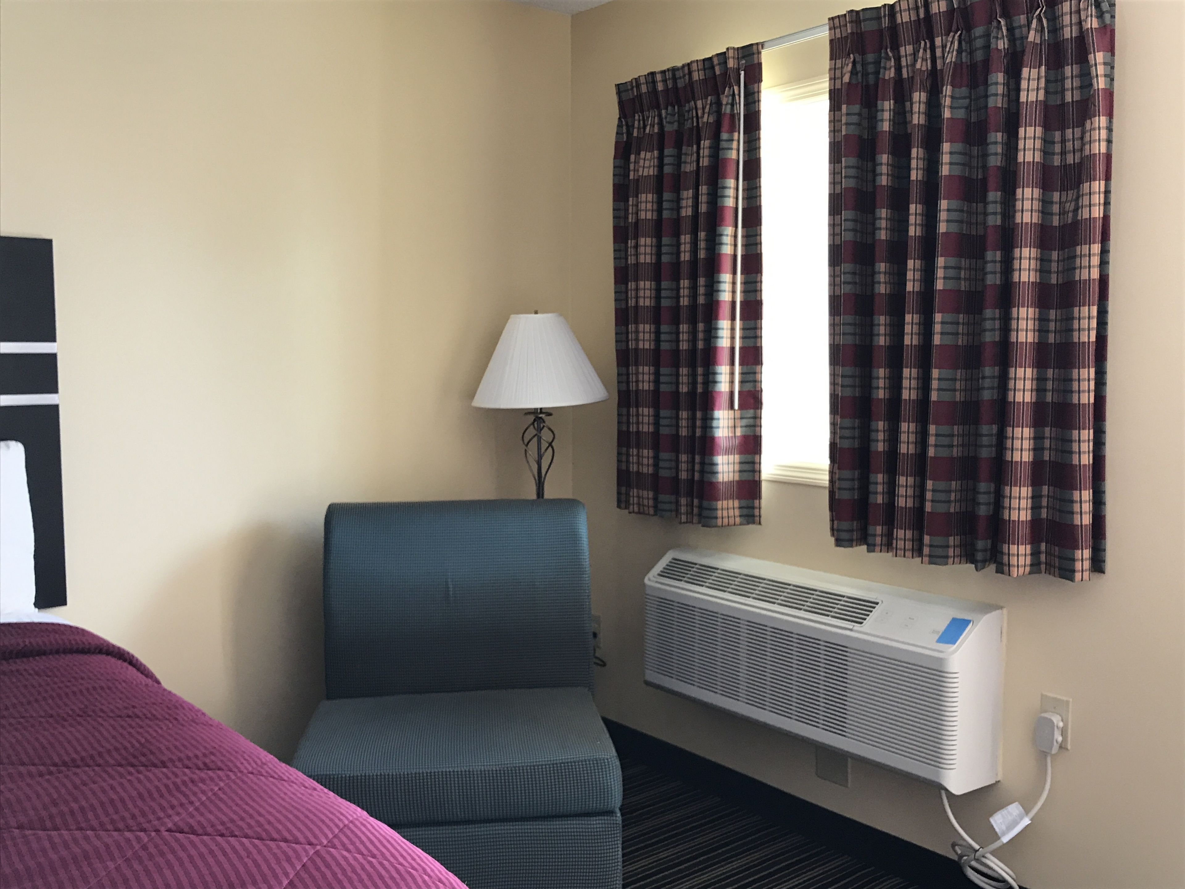 Ein Hotelzimmer mit Bett, Sessel, Lampe, einem Fenster mit Vorhängen und einer Klimaanlage.