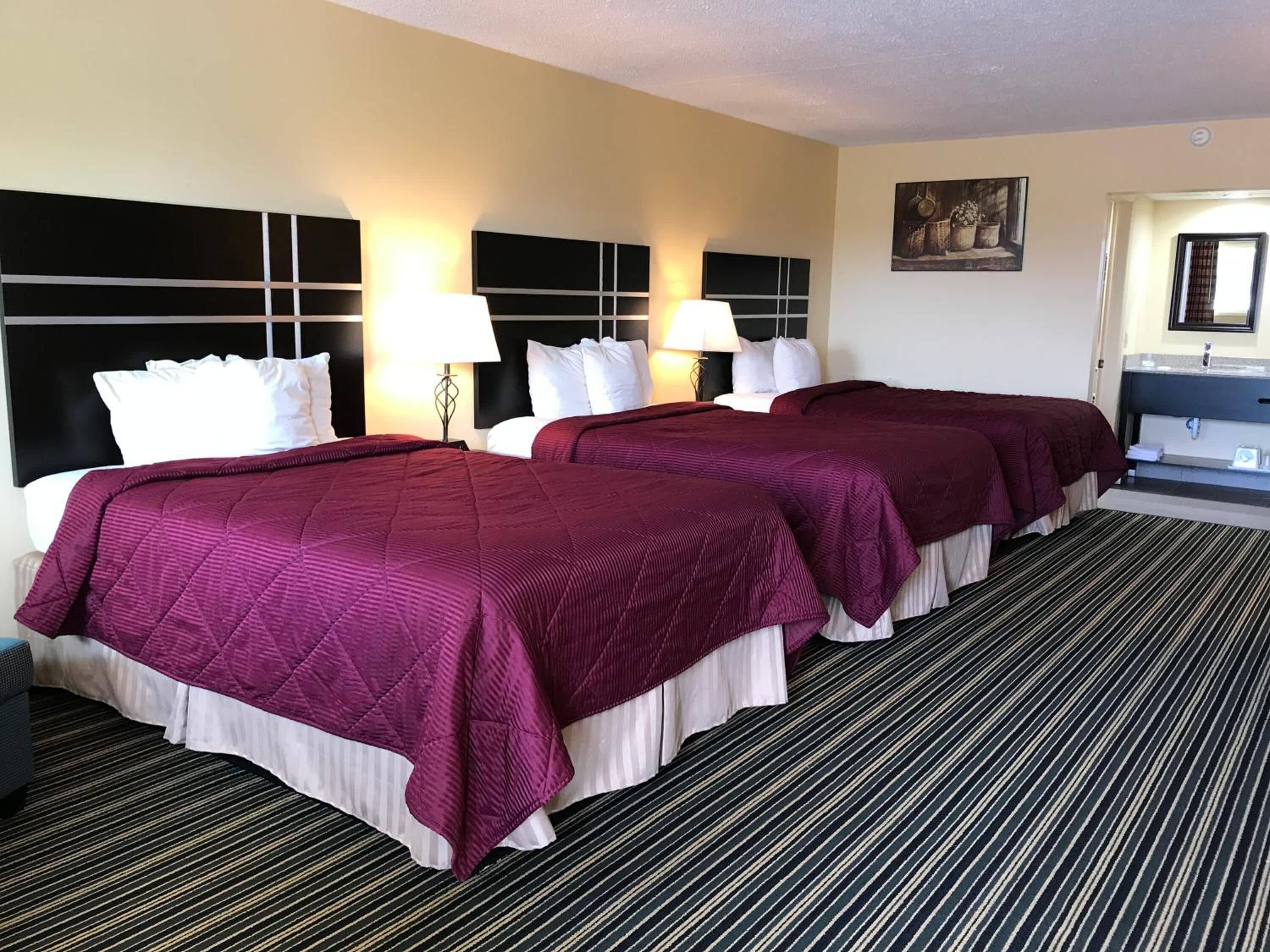 Une chambre d’hôtel avec deux lits, chacun doté d’une tête de lit, d’oreillers blancs et d’une couvre-lit bordeaux. On y trouve deux lampes de chevet, une petite table et une salle de bain visible à travers une porte ouverte.