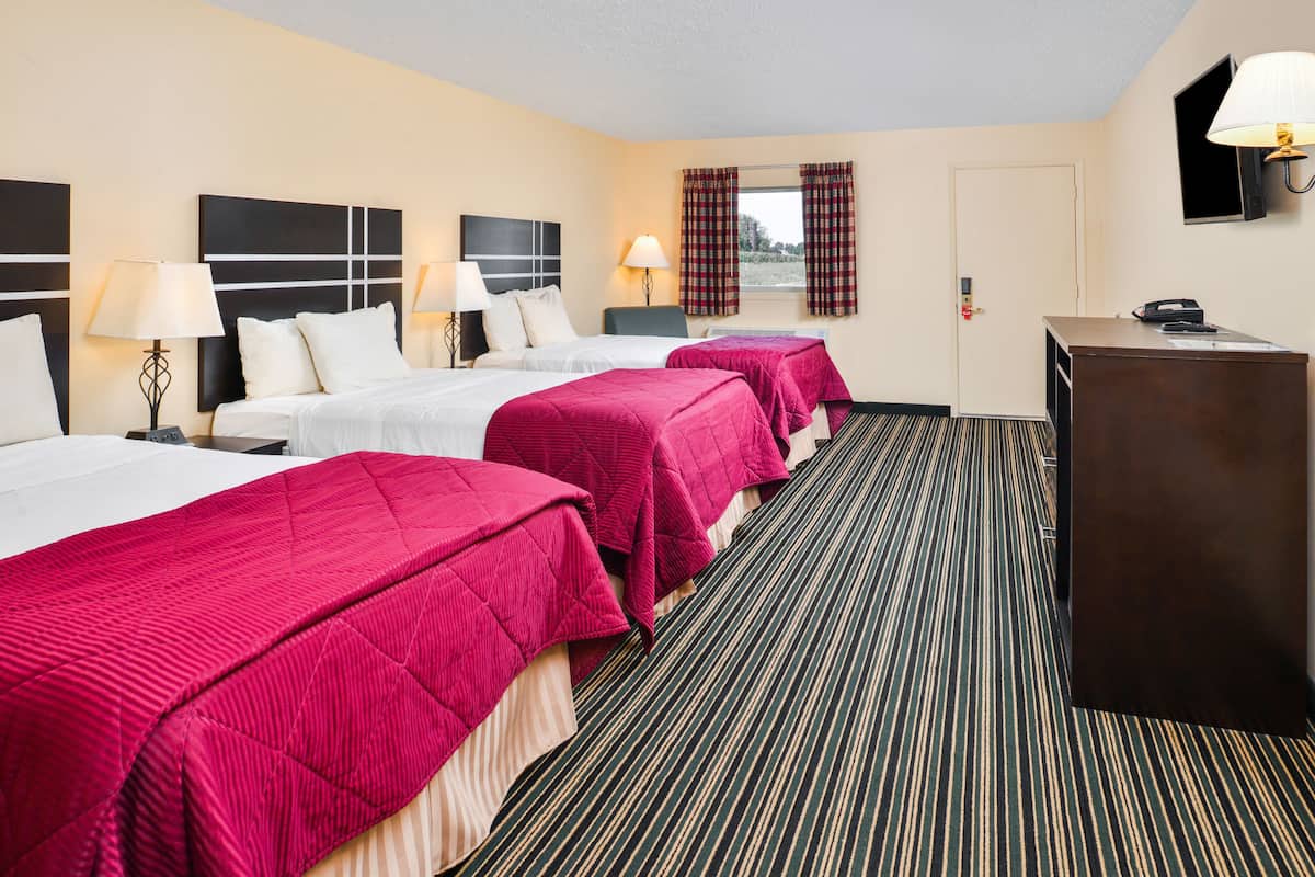 A hotel room with three beds, each with a red bedspread, a desk, and a television.