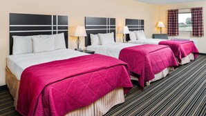 A hotel room with three beds, each with white linens and pink bedspreads. There are bedside tables with lamps, a window with curtains, and a carpeted floor.