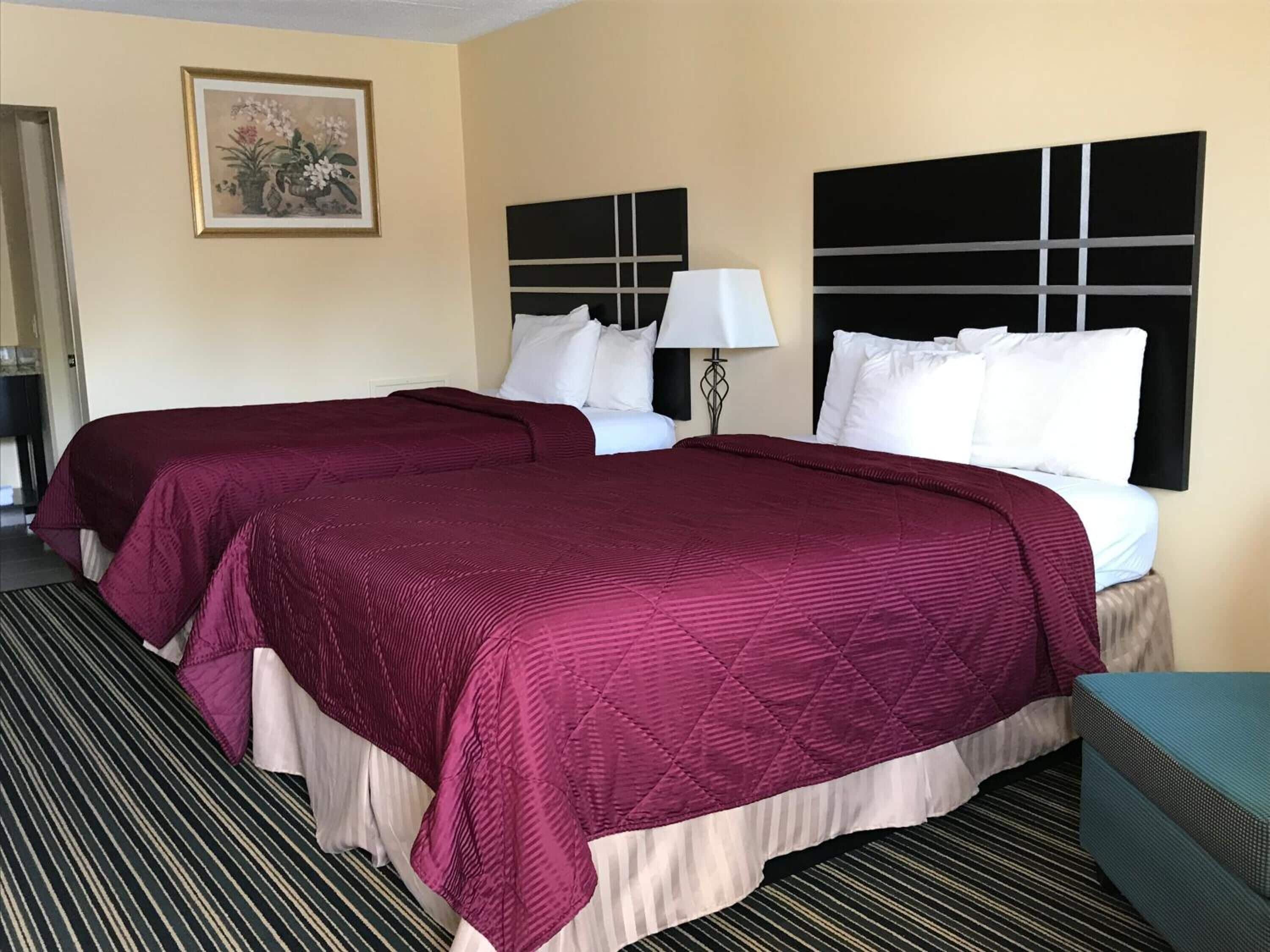 A hotel room with two beds, each with a red bedspread and white pillows, a black headboard, a bedside lamp, and a framed floral artwork on the wall.