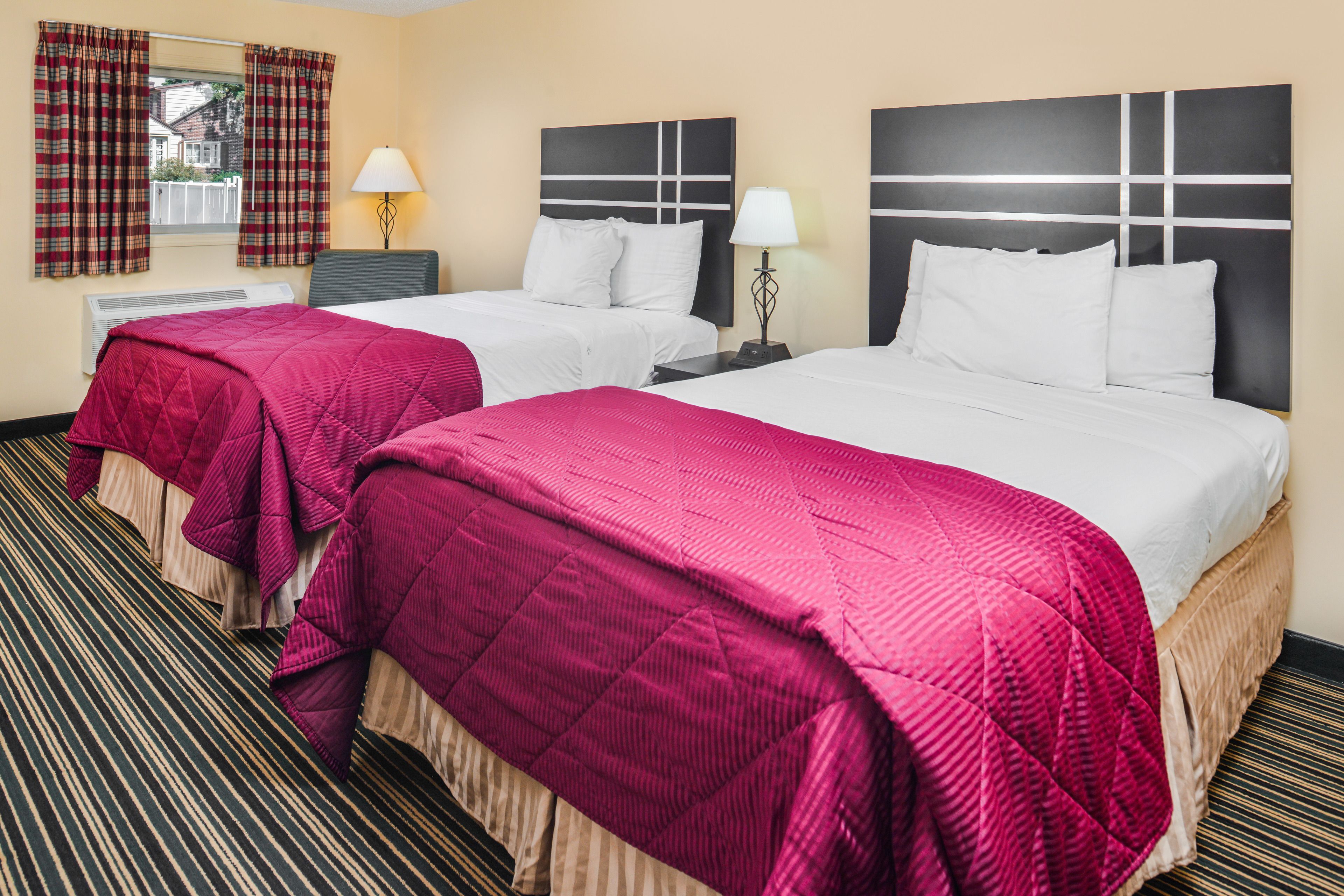 Une chambre d’hôtel avec deux lits, chacun doté d’une tête de lit blanche et d’une couvre-lit rouge. Il y a deux tables de chevet avec des lampes, et une fenêtre avec des rideaux à carreaux.