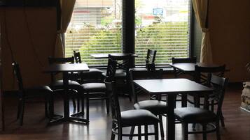 Interior de un restaurante con varias mesas y sillas, amplias ventanas con cortinas y vistas al exterior.