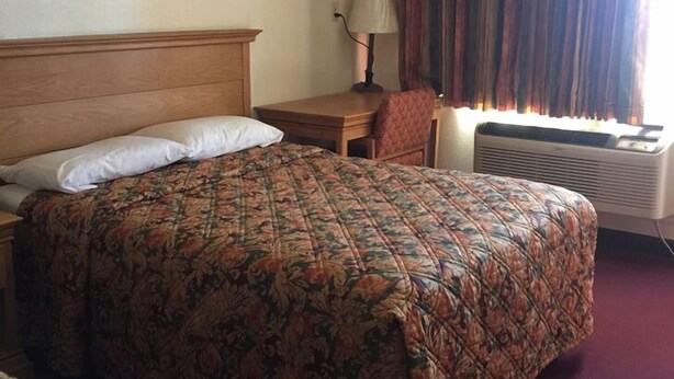 A hotel room with a large bed, a bedside table, a lamp, a chair, and an air conditioning unit. - Fabens Inn Motel (Fabens)