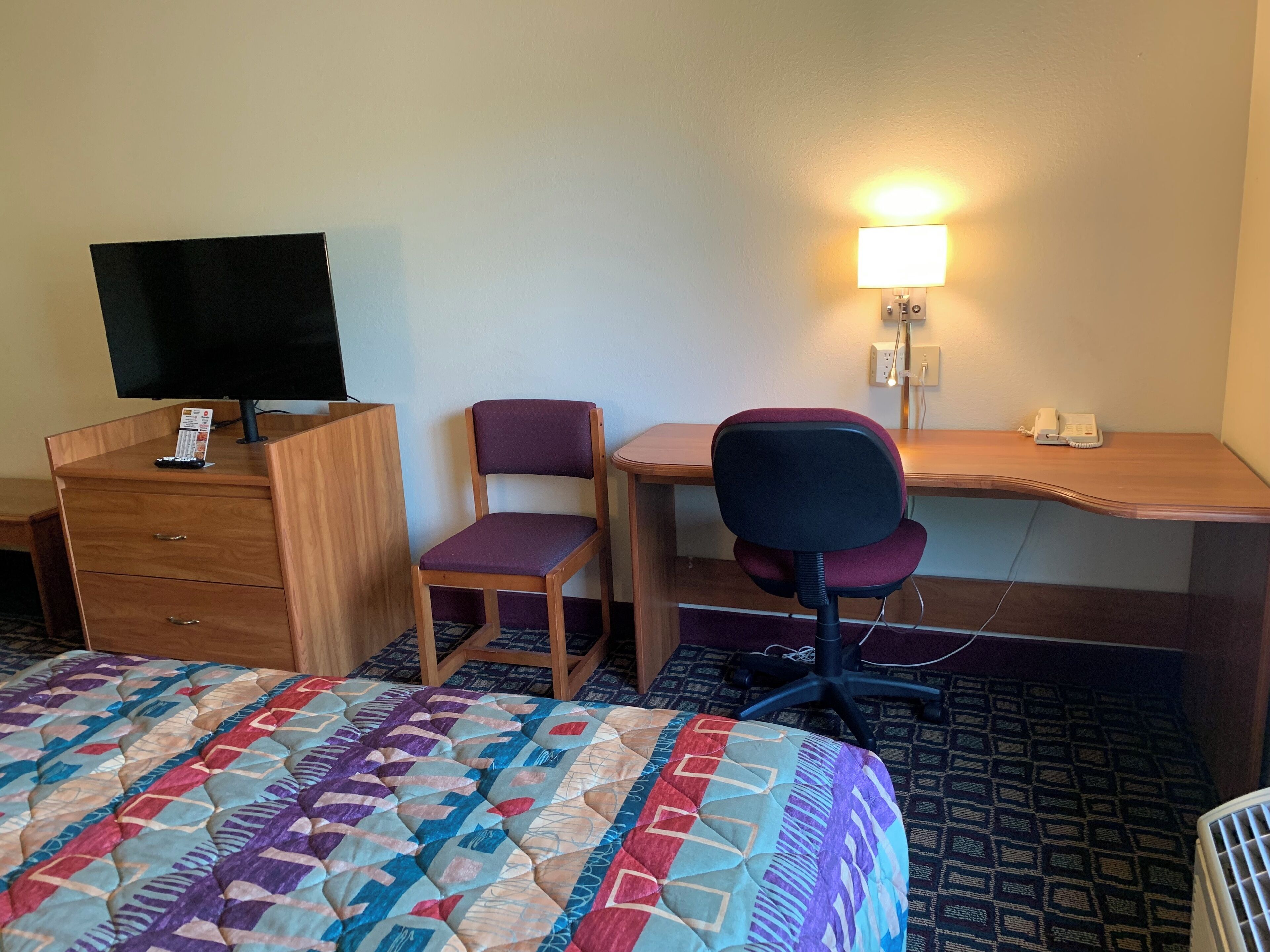 Une chambre d’hôtel avec un lit, un bureau en bois, un ordinateur, une chaise, une télévision et une lampe de chevet.