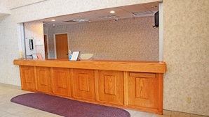 A reception desk with a wooden finish, a mirror above it, and a red doormat in front. - Budget Host Inn (Allegan)