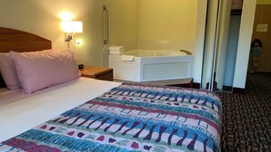 A hotel room with a bed, a bedside table, a lamp, a bathroom with a bathtub, and a patterned carpet. - Budget Host Inn (Allegan)