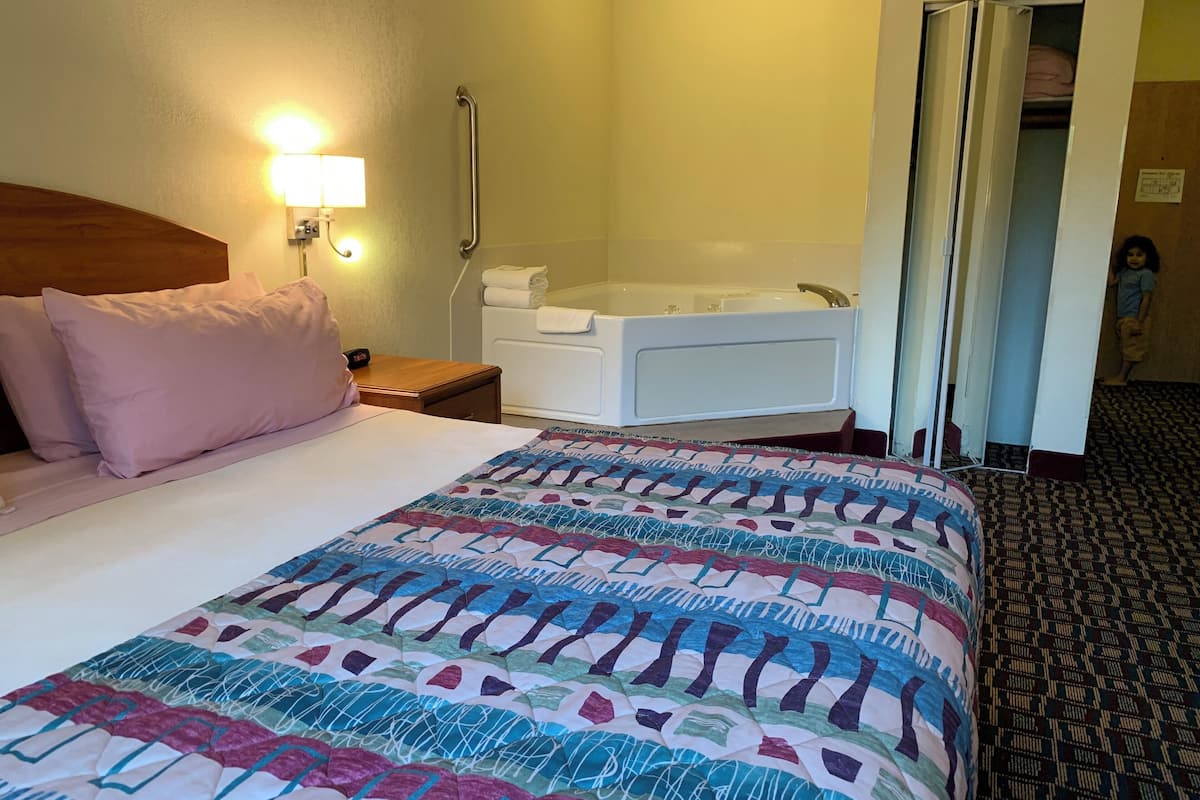 Une chambre d’hôtel comprenant un lit, une table de chevet, une lampe, une salle de bain avec baignoire et une moquette à motifs.
