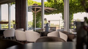 A restaurant interior with large windows, cushioned seating, and a view of greenery outside. - Van der Valk Hotel ARA (Zwijndrecht)
