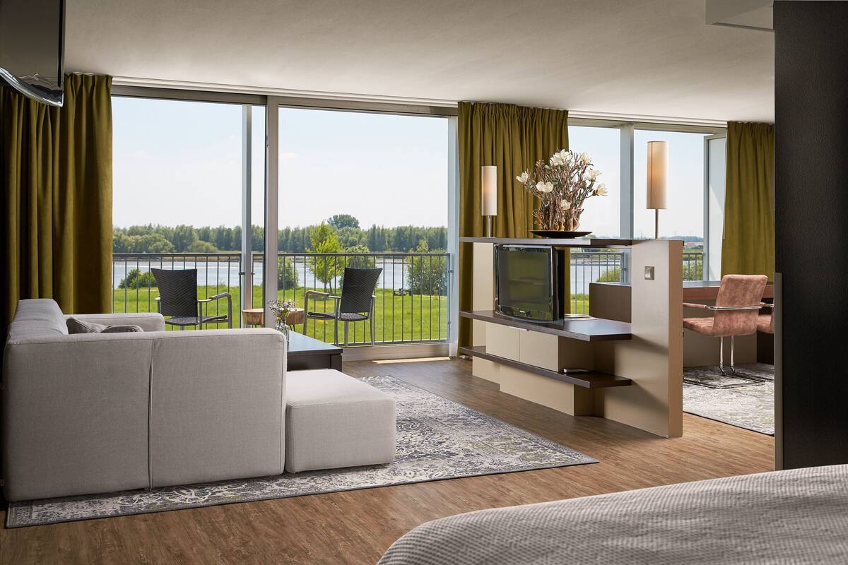 A modern hotel room with a large balcony, a sofa, a TV, and a dining area.