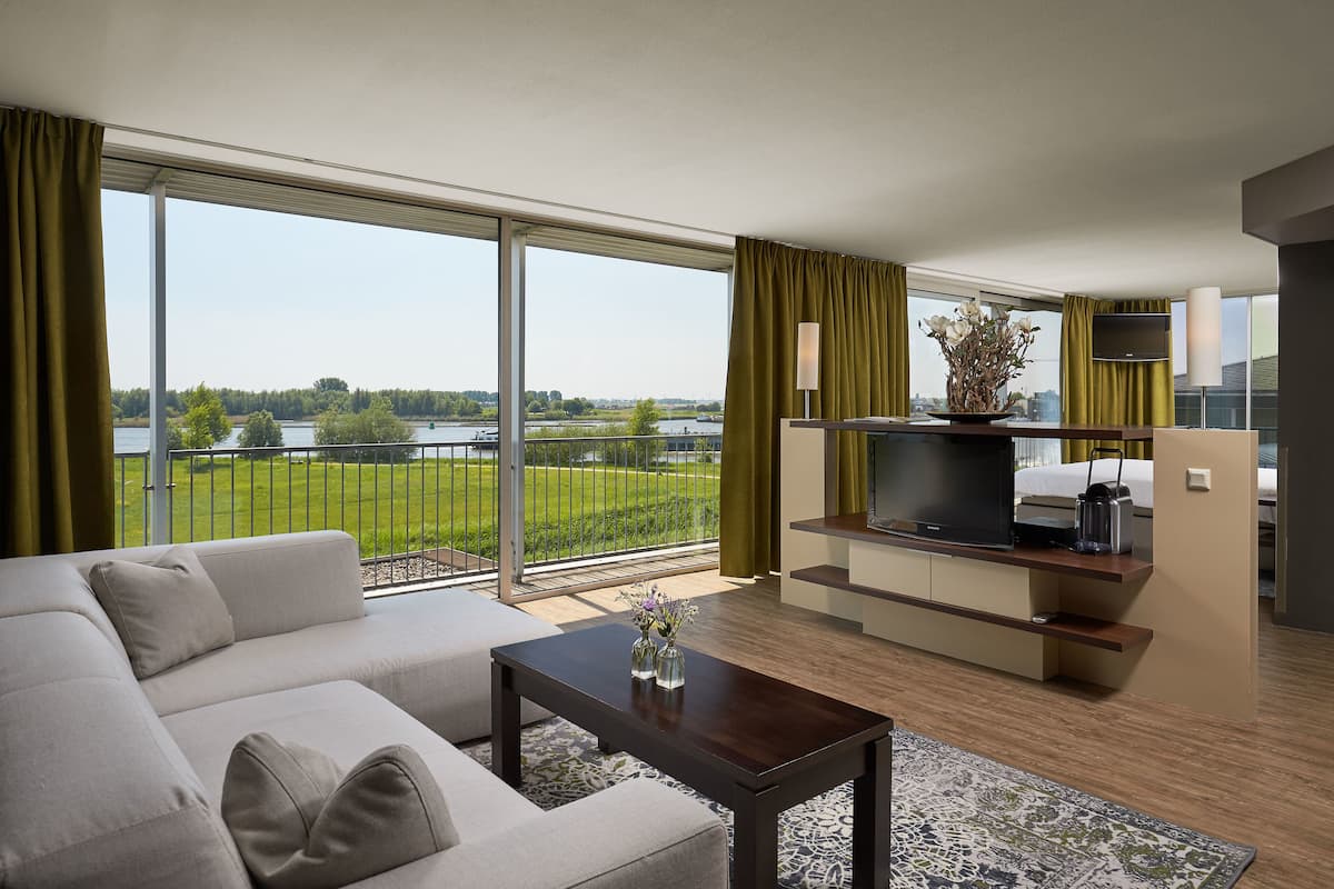 A modern living room with a sofa, coffee table, and TV stand, featuring a large window with a view of a lake and greenery.