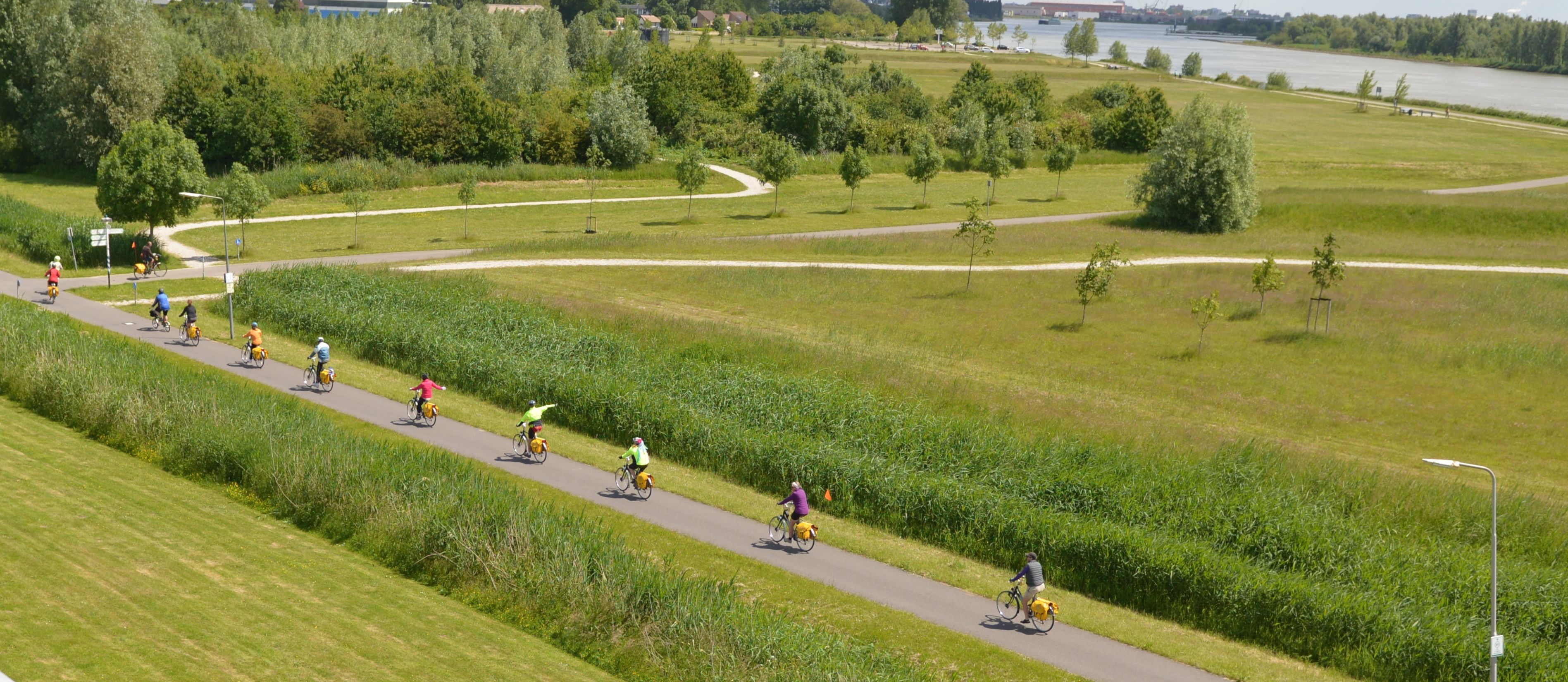 Une piste cyclable asphaltée, bordée de champs verdoyants et d’arbres, avec une rivière en arrière-plan, où l’on peut voir des cyclistes.