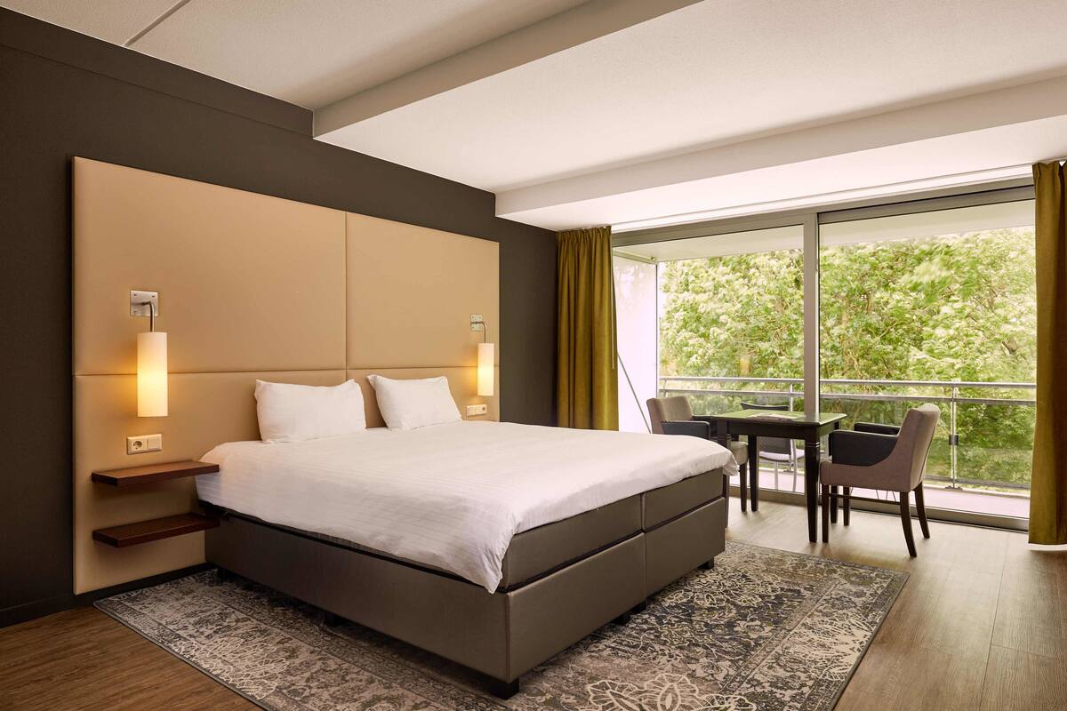 A modern hotel room with a large bed, bedside tables, a small table with chairs, and a balcony with a view of greenery.