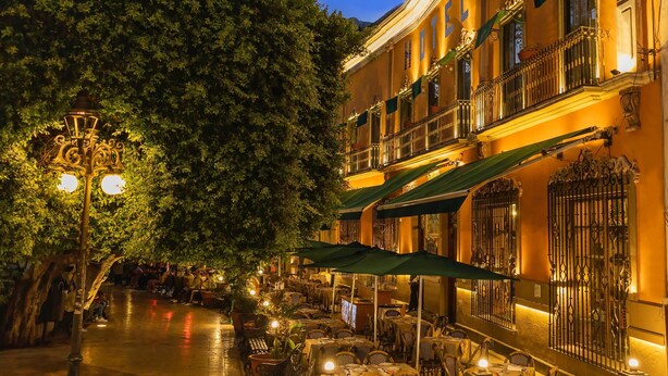 Un área de café al aire libre con mesas y sillas, un árbol y un edificio con balcón.