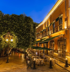 Un área de café al aire libre con mesas y sillas, un árbol y un edificio con balcón.