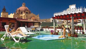 Un área de piscina en la azotea con sillones de descanso y una piscina pequeña, rodeada de plantas en macetas y un edificio con cúpula y paredes blancas.