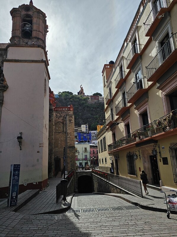 Una calle adoquinada con edificios históricos, una torre de campanas y una estatua en una colina.