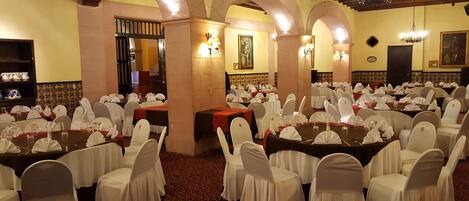 Un salón de banquetes con mesas redondas, sillas blancas y candelabros.