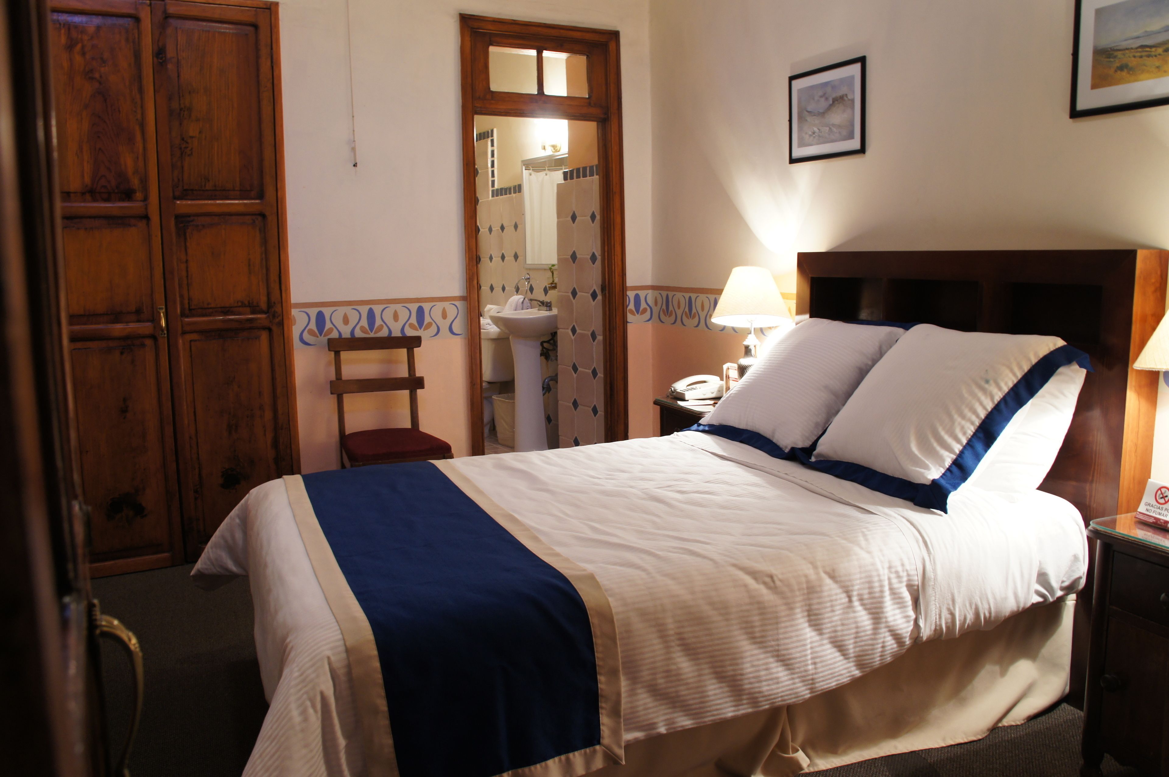 Une chambre d’hôtel avec un lit, une table de chevet, une lampe, une armoire en bois et une salle de bain visible à travers une porte ouverte.