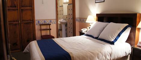 Habitación de hotel con cama, mesita de noche, lámpara, armario de madera y baño visible a través de una puerta abierta.