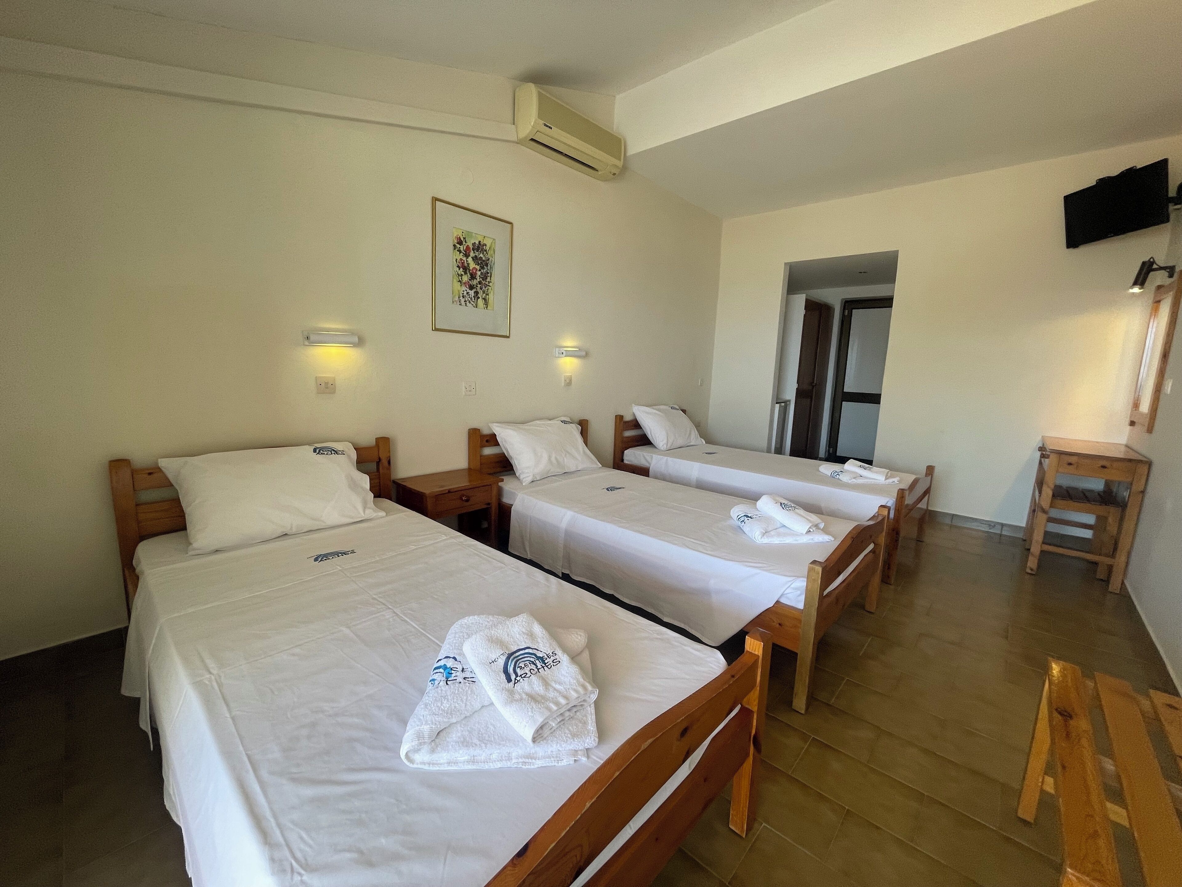 A hotel room with three single beds, each with a white bedspread and a towel with a logo. There is a wooden nightstand with a lamp on each bed, a wall-mounted air conditioner, and a framed picture above the beds.