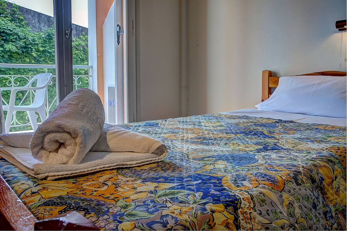 A bed with a floral bedspread, a rolled towel, and a view of greenery outside.