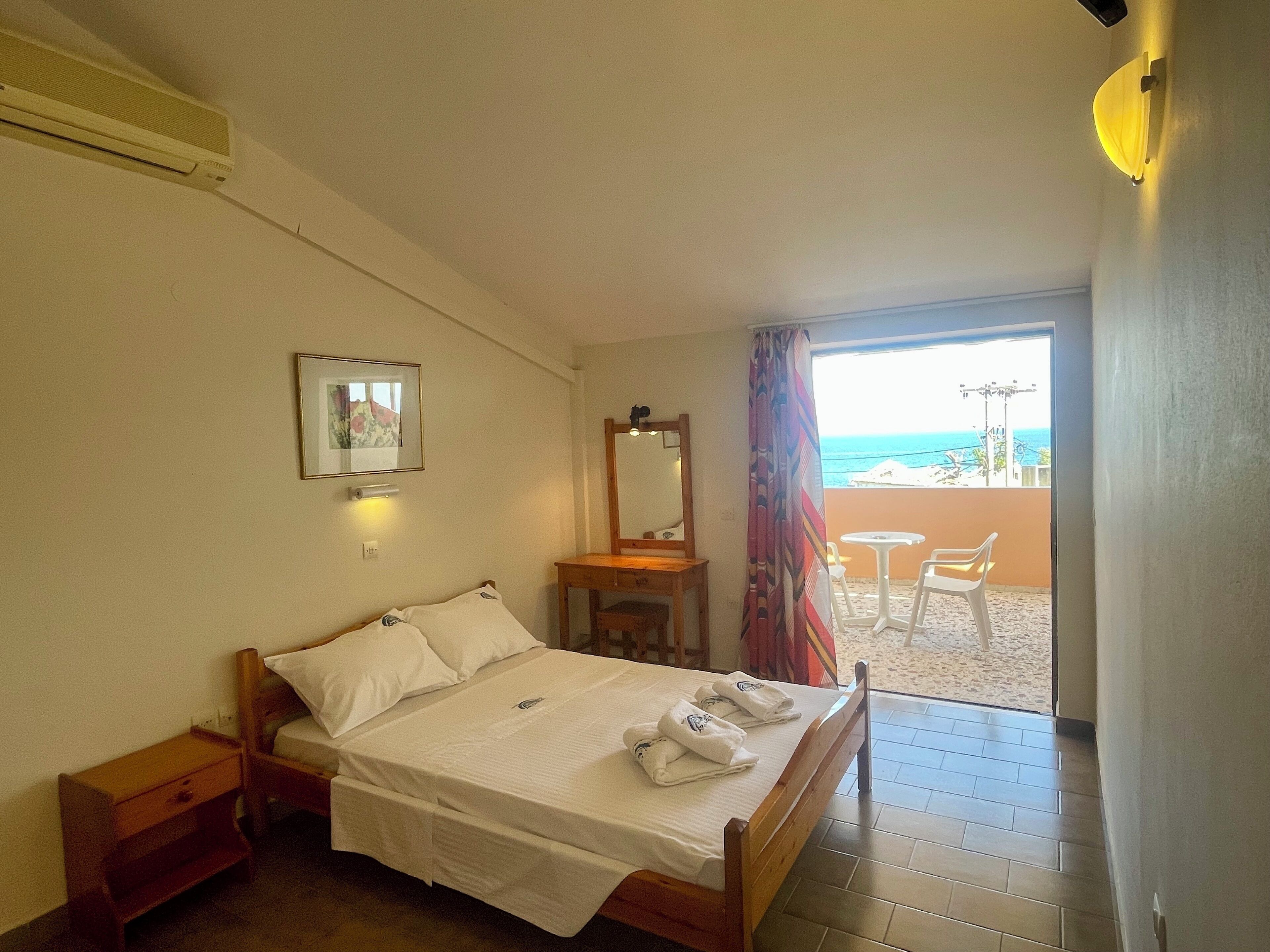 A hotel room with a bed, a bedside table, a mirror, a window with a view of the beach, and a balcony with chairs.