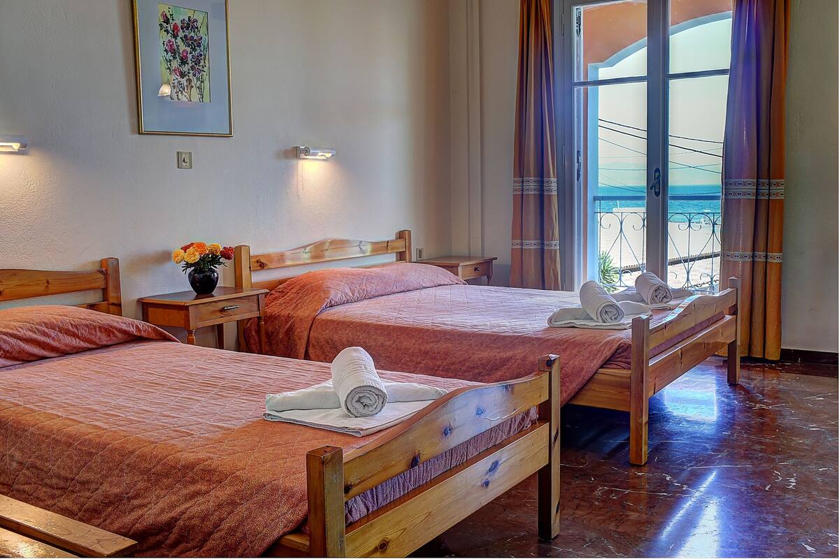 A hotel room with two wooden beds, a nightstand with a vase of flowers, and a window with curtains.