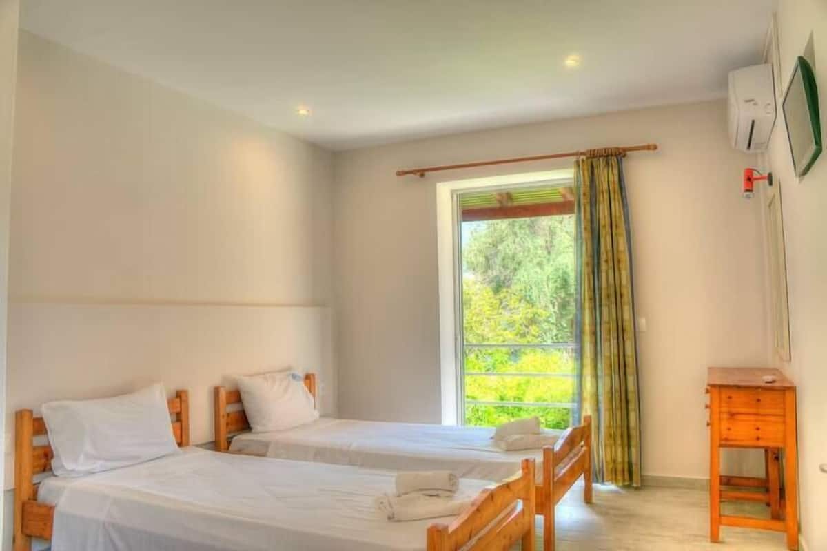 A room with two wooden beds, a small wooden table, and a window with a view of greenery.