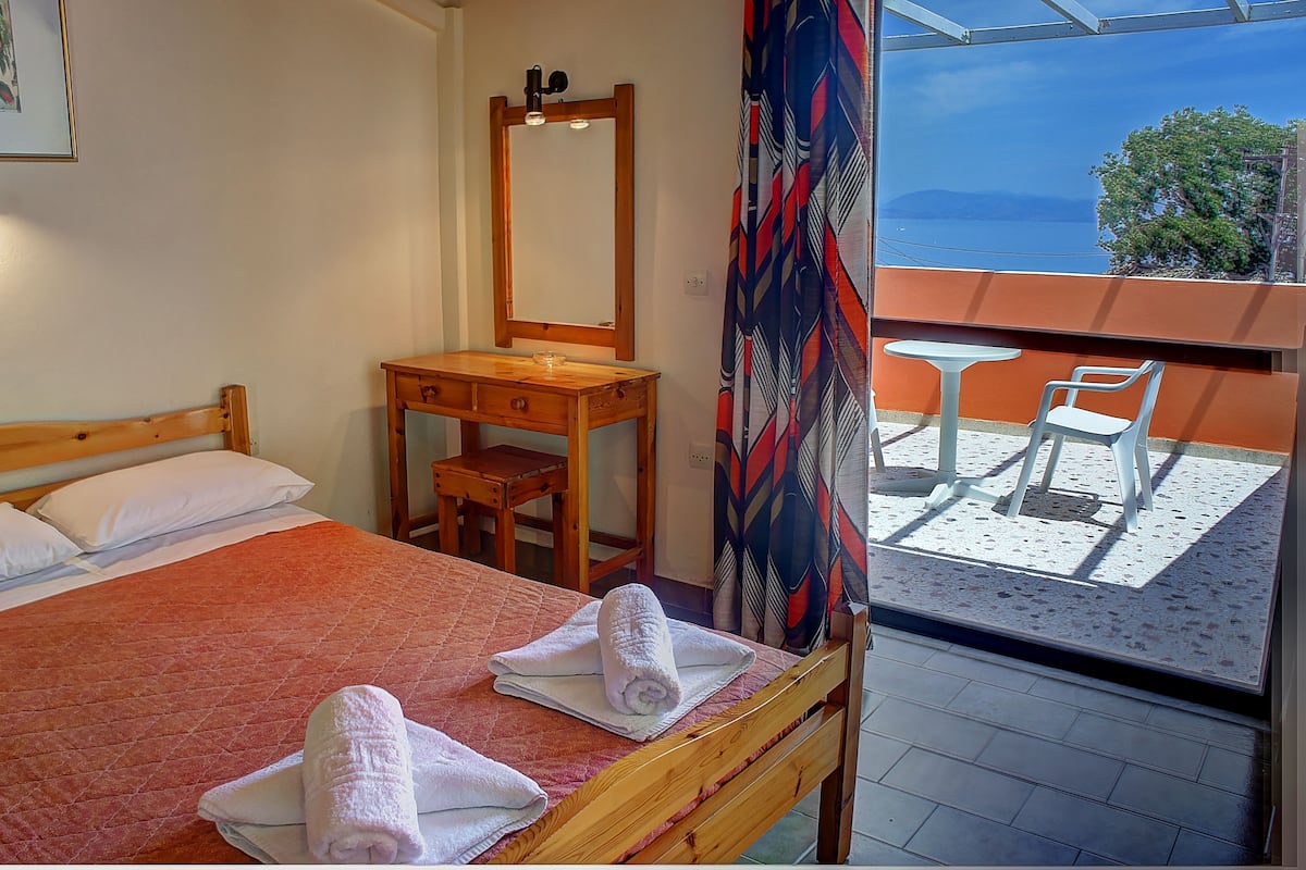 A hotel room with a bed, a wooden dresser, a mirror, and a balcony with a view of the sea.