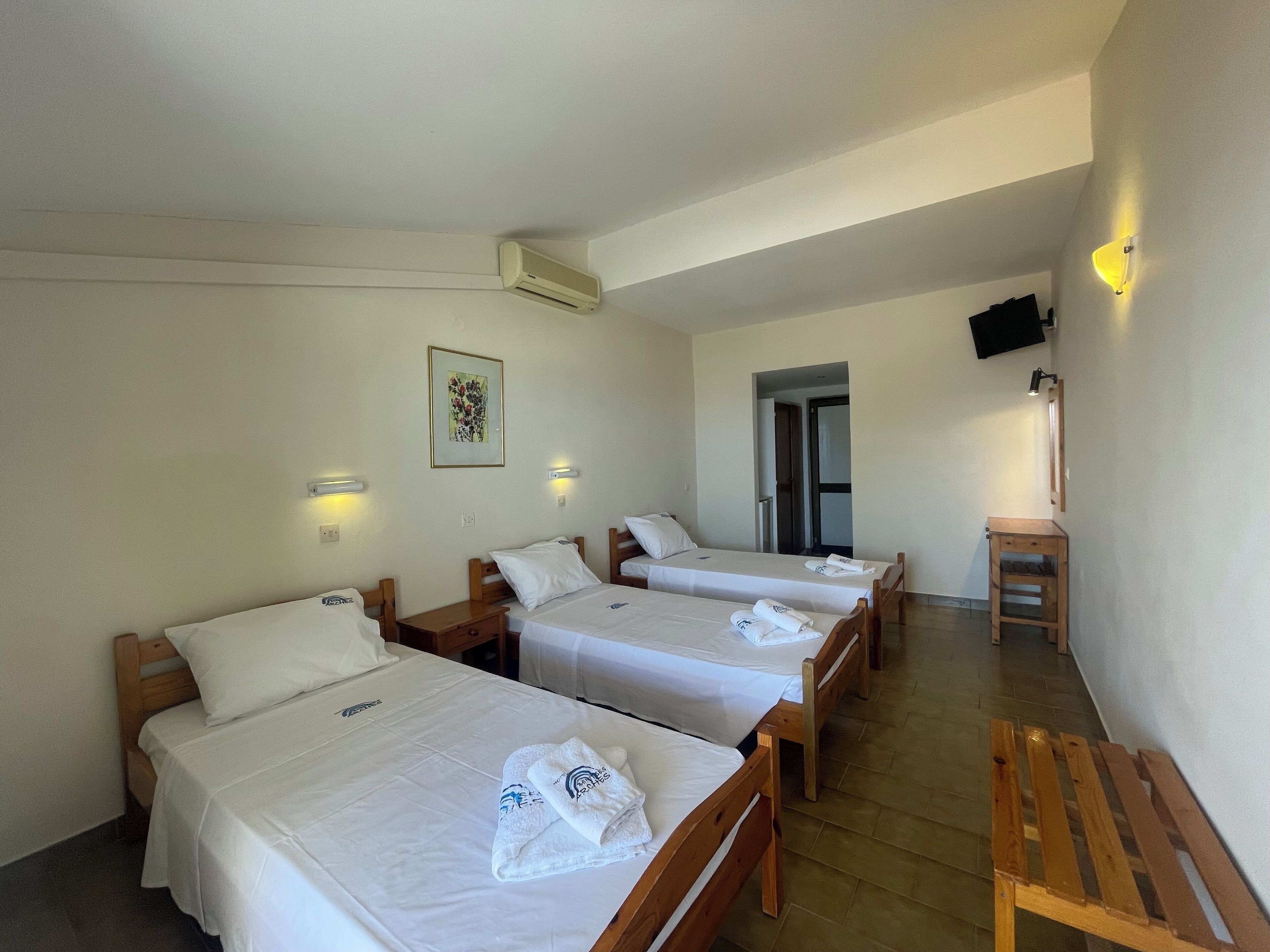 Habitación de hotel con tres camas individuales, un banco de madera, una mesita pequeña y un televisor fijado a la pared.