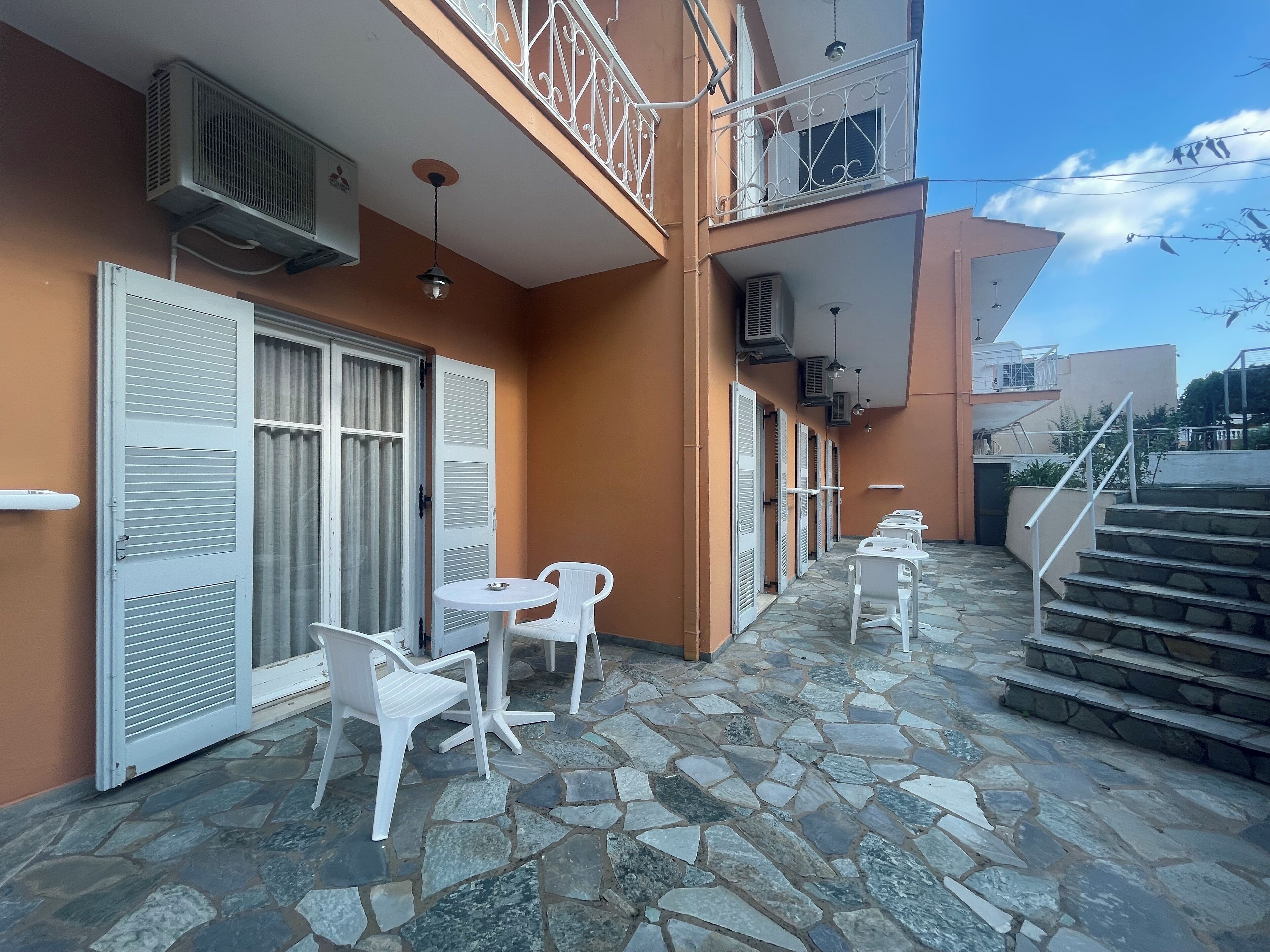 Un edificio con patio de losas de piedra, sillas blancas y una escalera que conduce a un nivel superior.