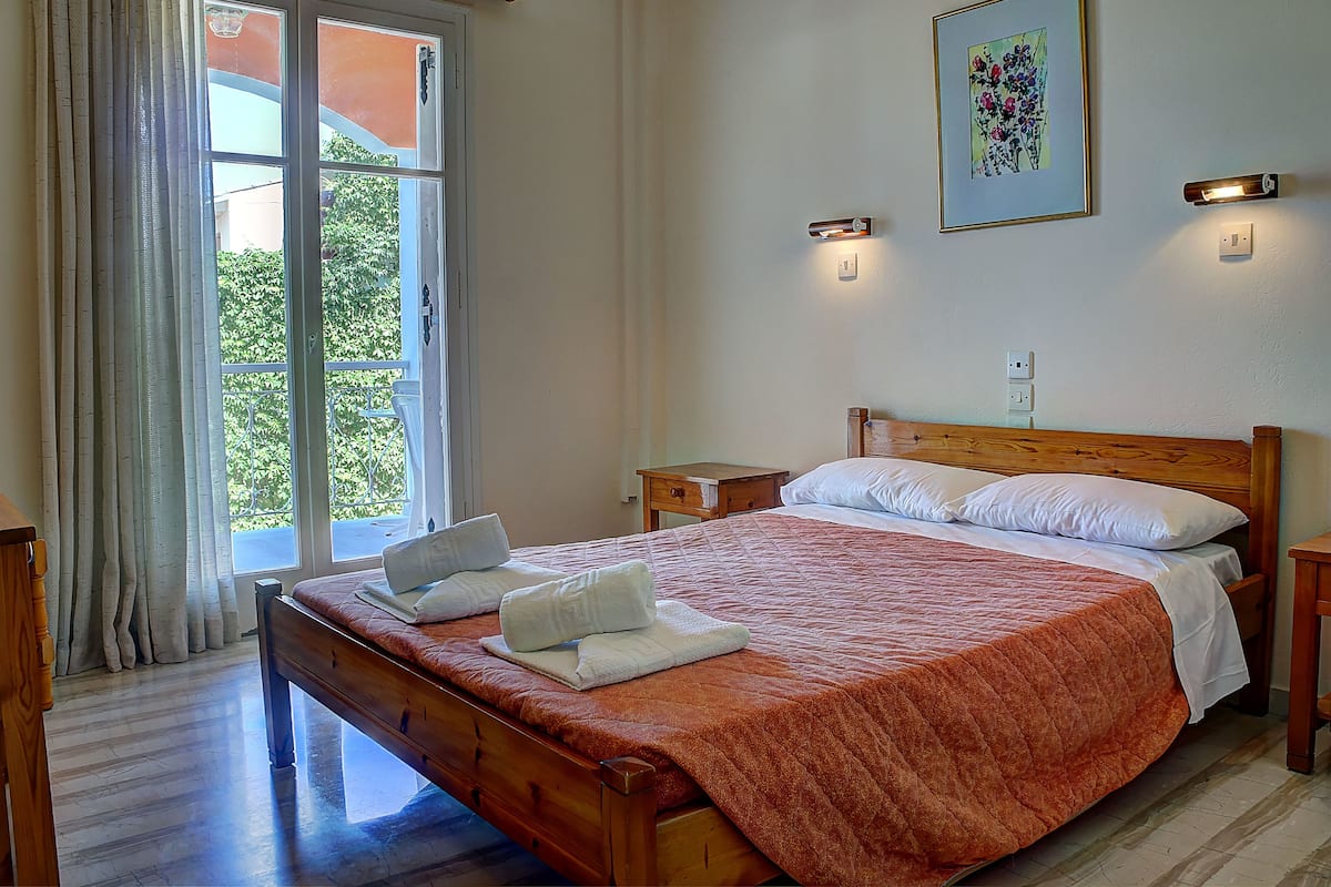 A neatly made bed with a red blanket, a wooden headboard, and white pillows. There is a window with curtains, a bedside table, and a wall-mounted light fixture.