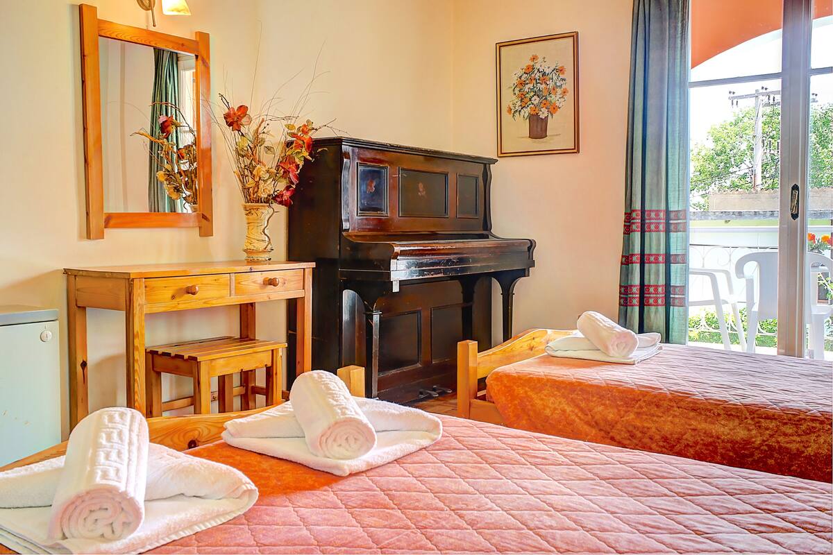 A hotel room with two beds, a piano, a mirror, a vase with flowers, and a view of the outdoors.