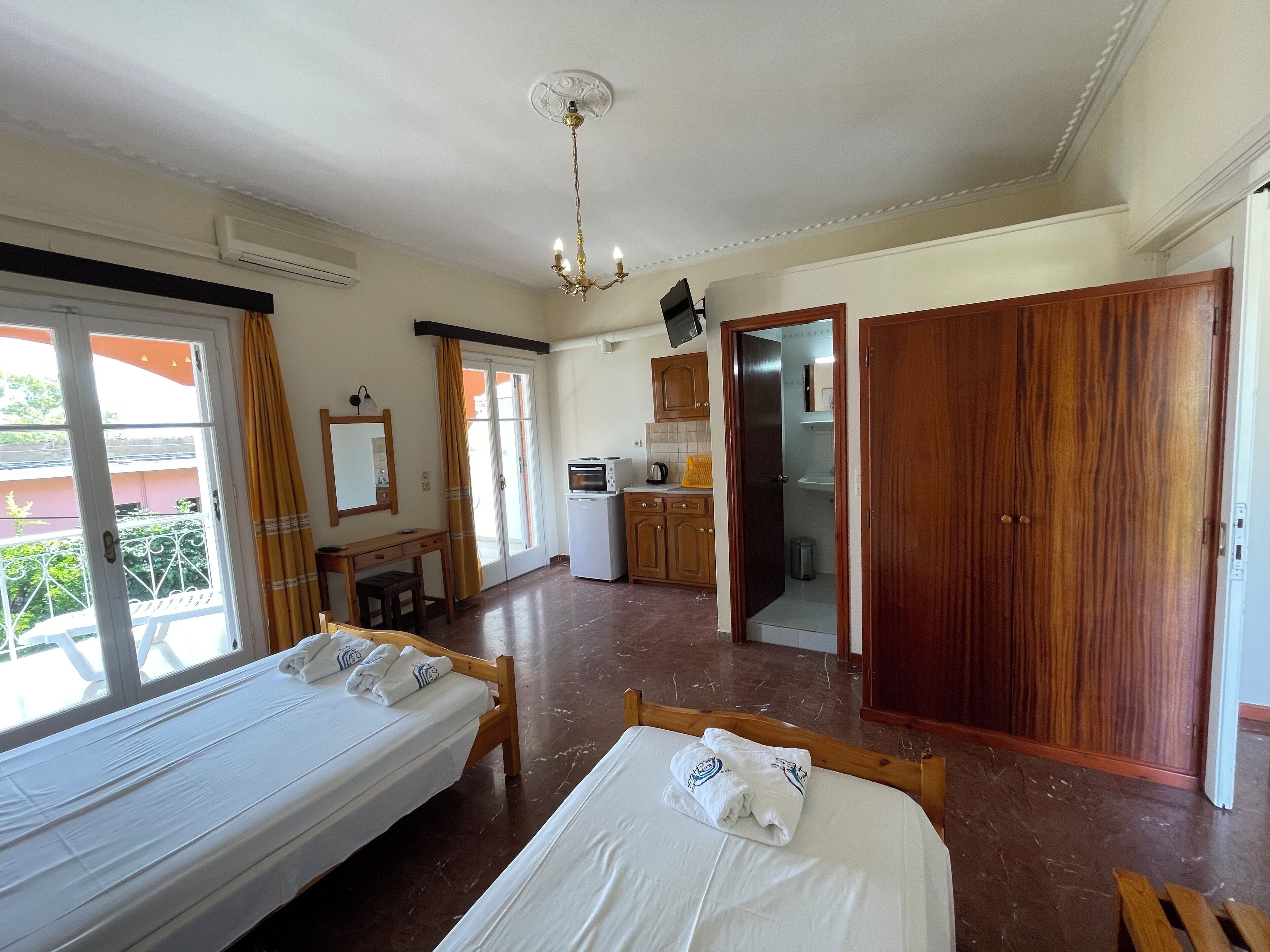 A hotel room with two beds, a kitchenette, a bathroom, and a view of the outside balcony.
