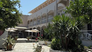 A courtyard with a stone floor, potted plants, outdoor seating, and a flag on a building. - Benitses Arches (Corfu)