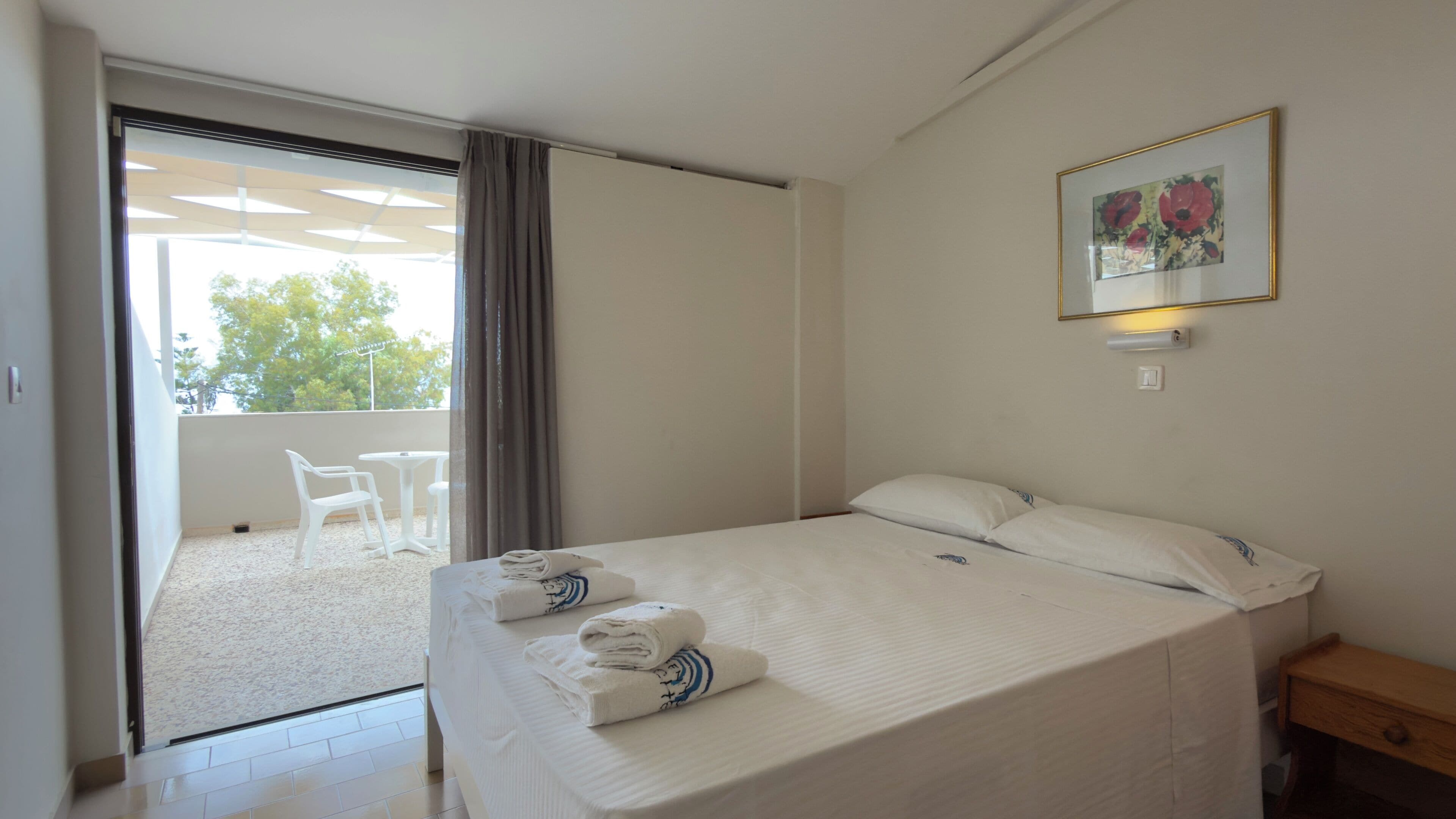 A hotel room with a bed, a small wooden nightstand, a wall-mounted light, a framed picture, and a balcony with white chairs and a table.