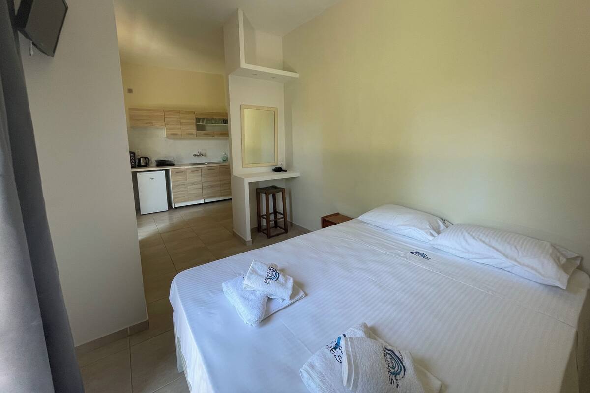 A hotel room with a bed, two towels, a small table, a nightstand, and a view of the kitchen.