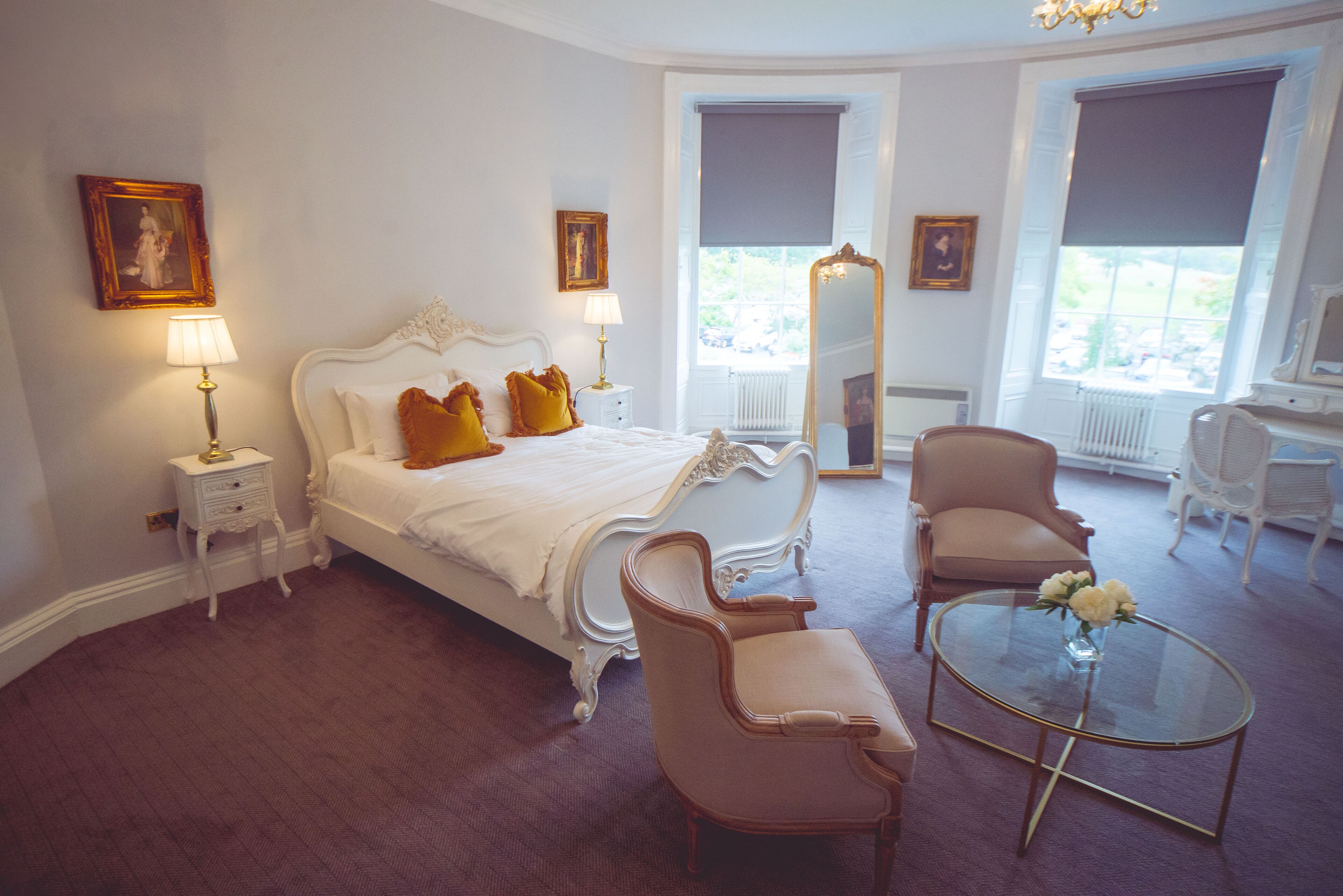 A bedroom with a large bed, two chairs, a round glass table with flowers, and a large mirror.