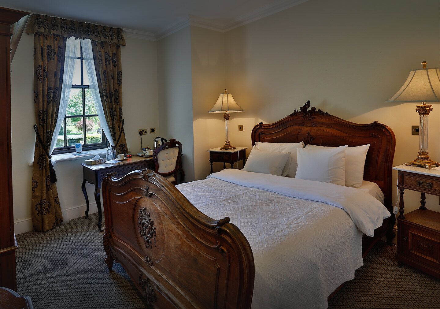 A classic wooden bed with a white bedspread, a nightstand with a lamp, a desk with a chair, and a window with curtains.