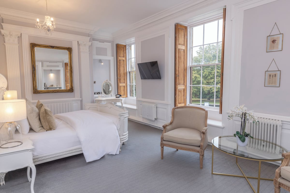 Un dormitorio clásico y elegantemente amoblado con una cama grande, un sillón, una mesita de vidrio y un televisor montado en la pared.