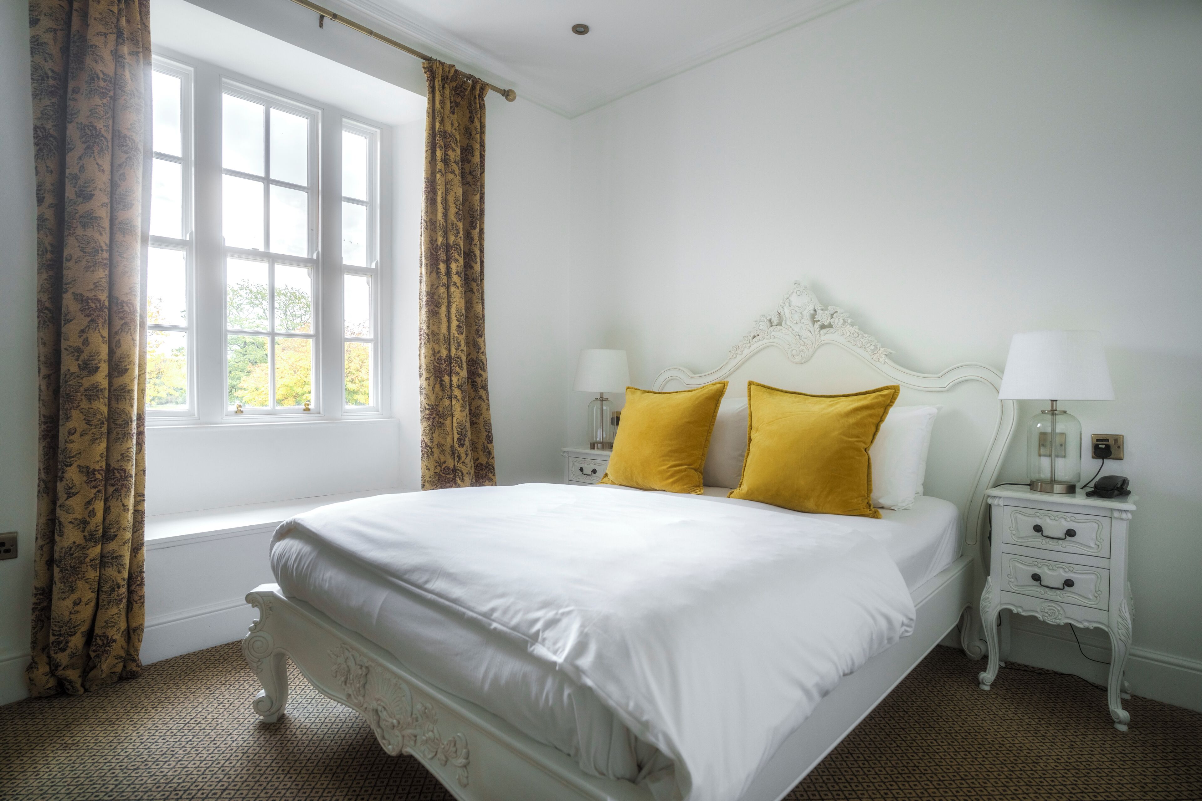 A white four-poster bed with yellow pillows, a white nightstand with a lamp, and a window with curtains.