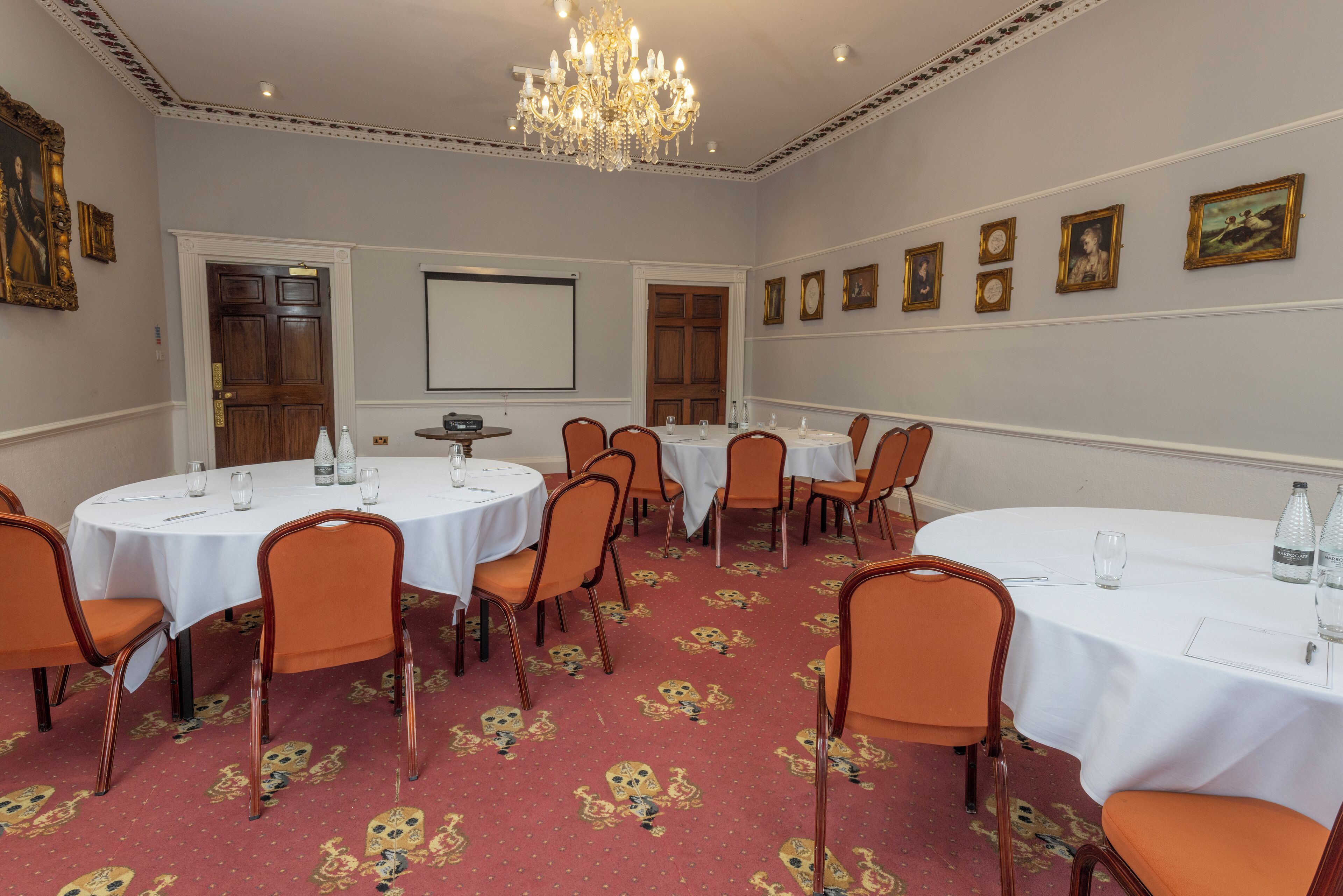 A conference room with round tables, orange chairs, a projector screen, and framed pictures on the wall.