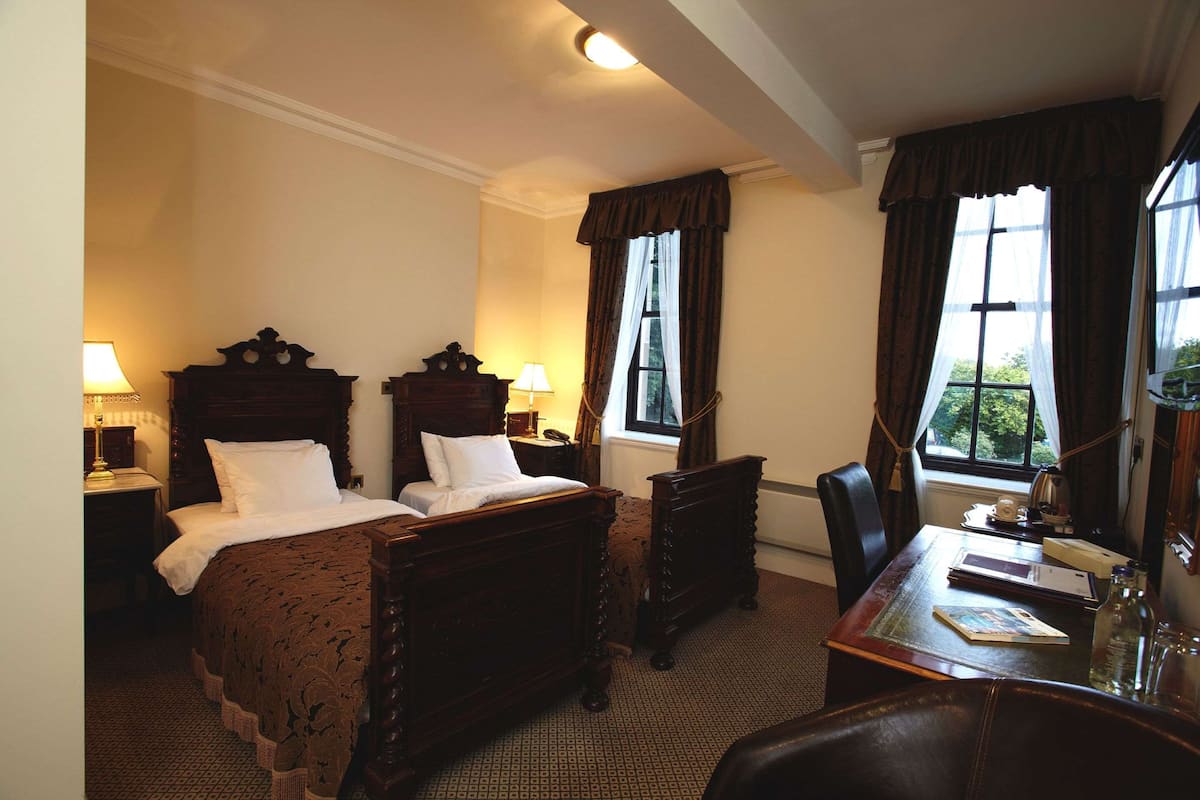 Habitación de hotel con dos camas de madera, un escritorio con silla, una lámpara y una ventana con cortinas.