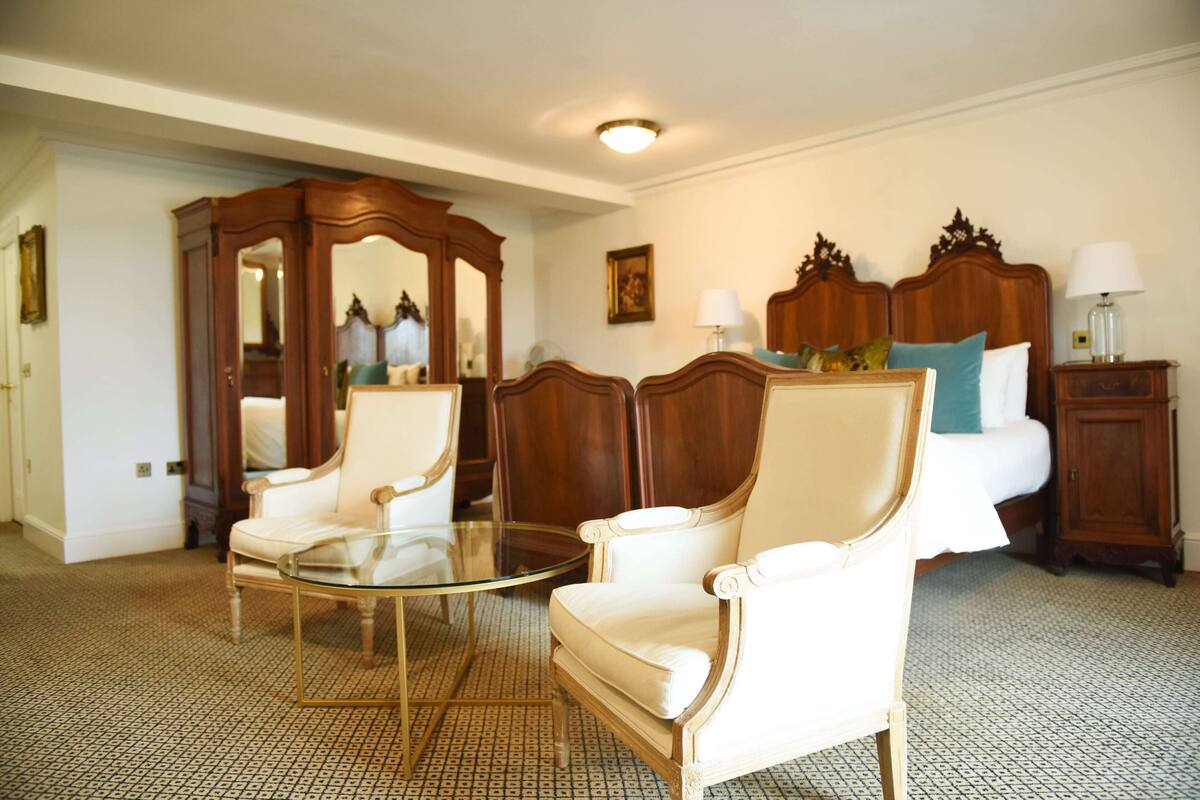 Habitación de hotel con cama, dos sillones, una mesa de centro de cristal, un armario de madera y una mesita de noche con lámpara.