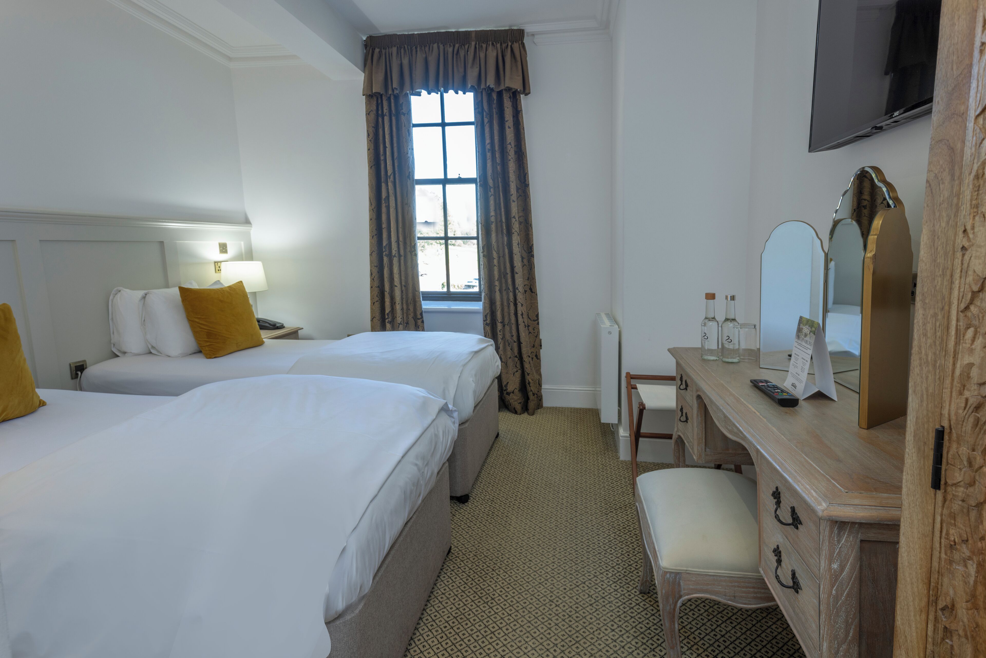 Habitación de hotel con dos camas, un aparador de madera, un espejo, un televisor y una ventana con cortinas.