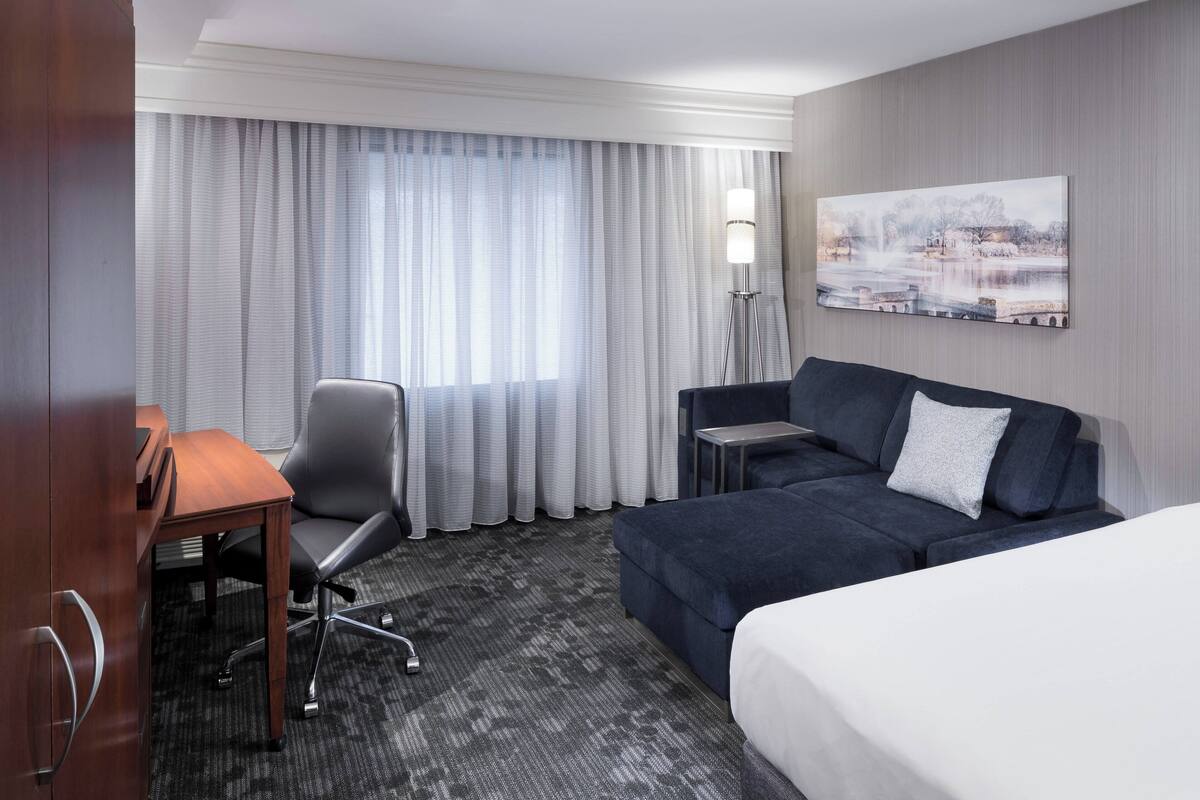 A hotel room with a bed, a desk, a chair, a sofa, a lamp, and a painting on the wall.