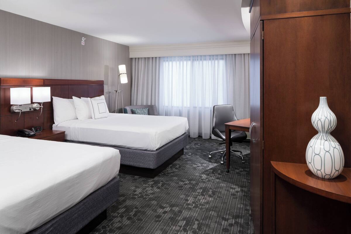 A hotel room with two beds, a desk, a chair, a lamp, and a vase on a side table.