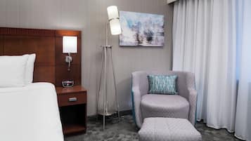 A hotel room with a bed, a desk with a lamp, a chair, a small table, and a painting on the wall.