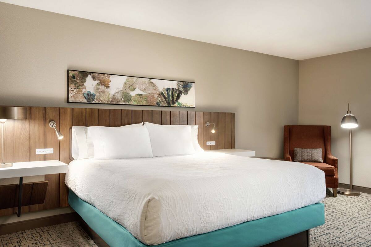 Ein Hotelzimmer mit einem großen Bett, einem Holzkopfteil, einem Wandbild, einem Schreibtisch mit Lampe und einem Sessel.